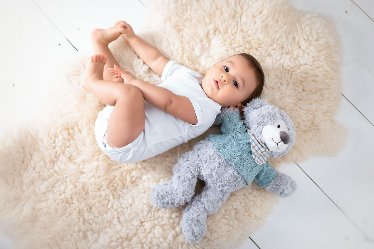 Ainda olhando para cima, o Eduardo está deitado em um tapetinho de carneiro. Ele segura os pezinhos e está com o seu urso deitado ao lado