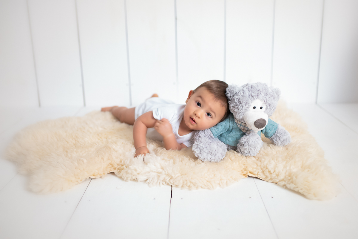 O Edu está de bruços, apoiado sob um dos seus cotovelos. Ele quase cai para o lado e encosta no ursinho que está sentado ao lado dele