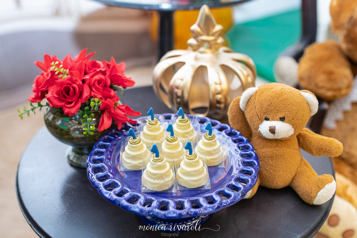 Mesa redonda, em cima aparador azul com docinhos em forma de bolo, do lado esquerdo um vaso com rosas, ao lado direito um urso e atrás uma coroa dourada 