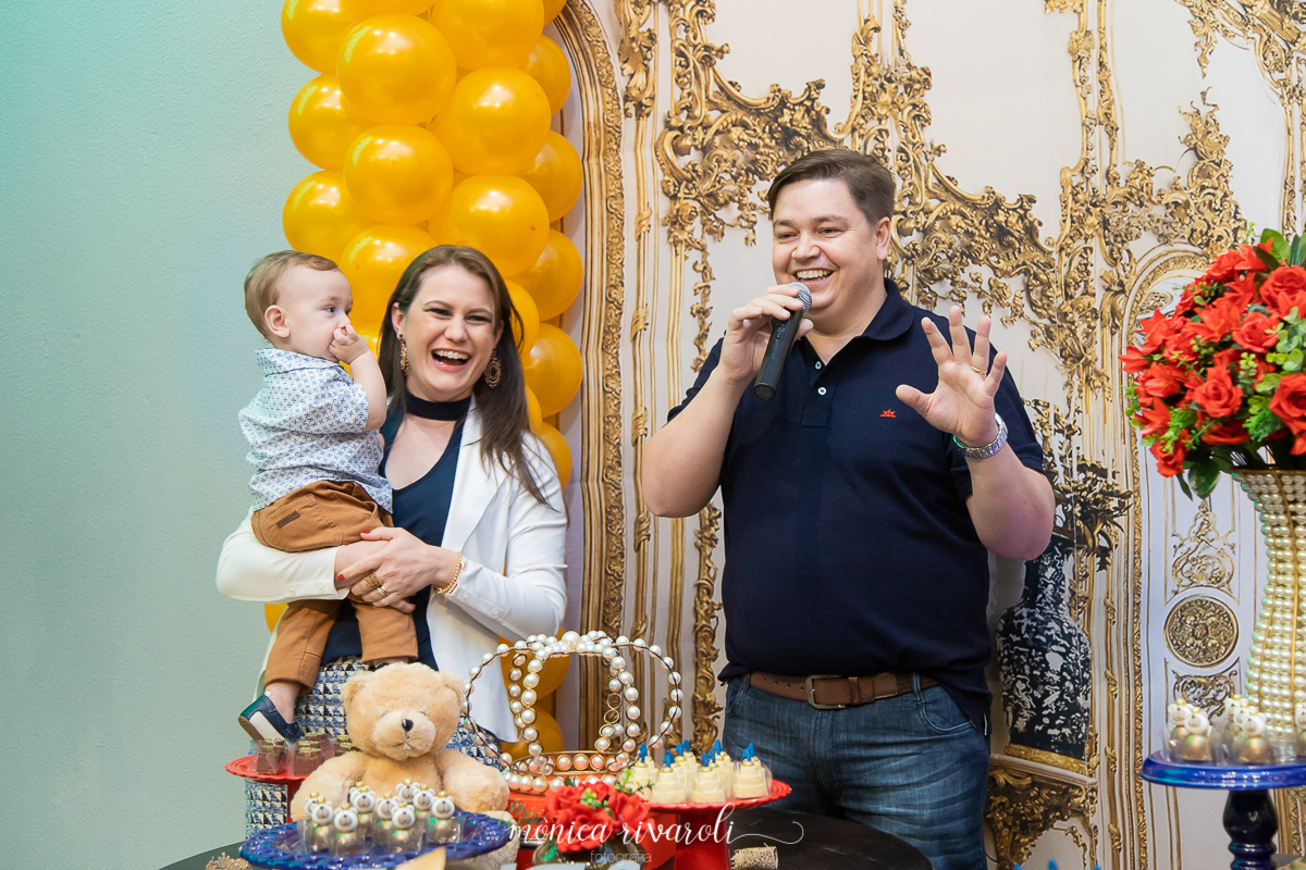 Papai falando no microfone, ao lado a mamãe que sorri com o Miguel no colo