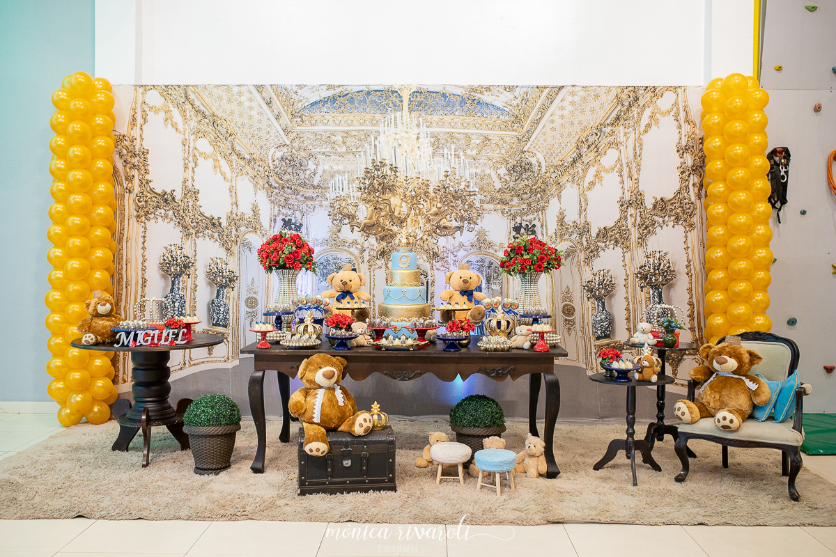 Imagem de toda a decoração de reinado. Com uma mesa grande no meio, ao lado esquerdo uma mesinha redonda e a direita duas mesinhas redondas pequenas e uma cadeira. 