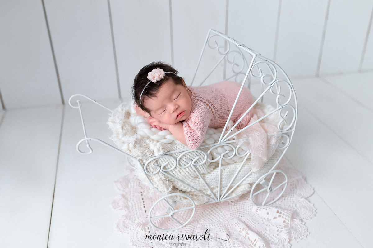 Em um carrinho de ferro todo trabalhada, a bebê dorme de bruços. O fundo da foto é de madeira branca e a bebê veste um macacão rosa