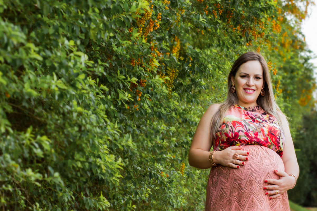 A mamãe está em fundo de folhas verdes com as mãos sobre a barriga com um sorriso lindo!