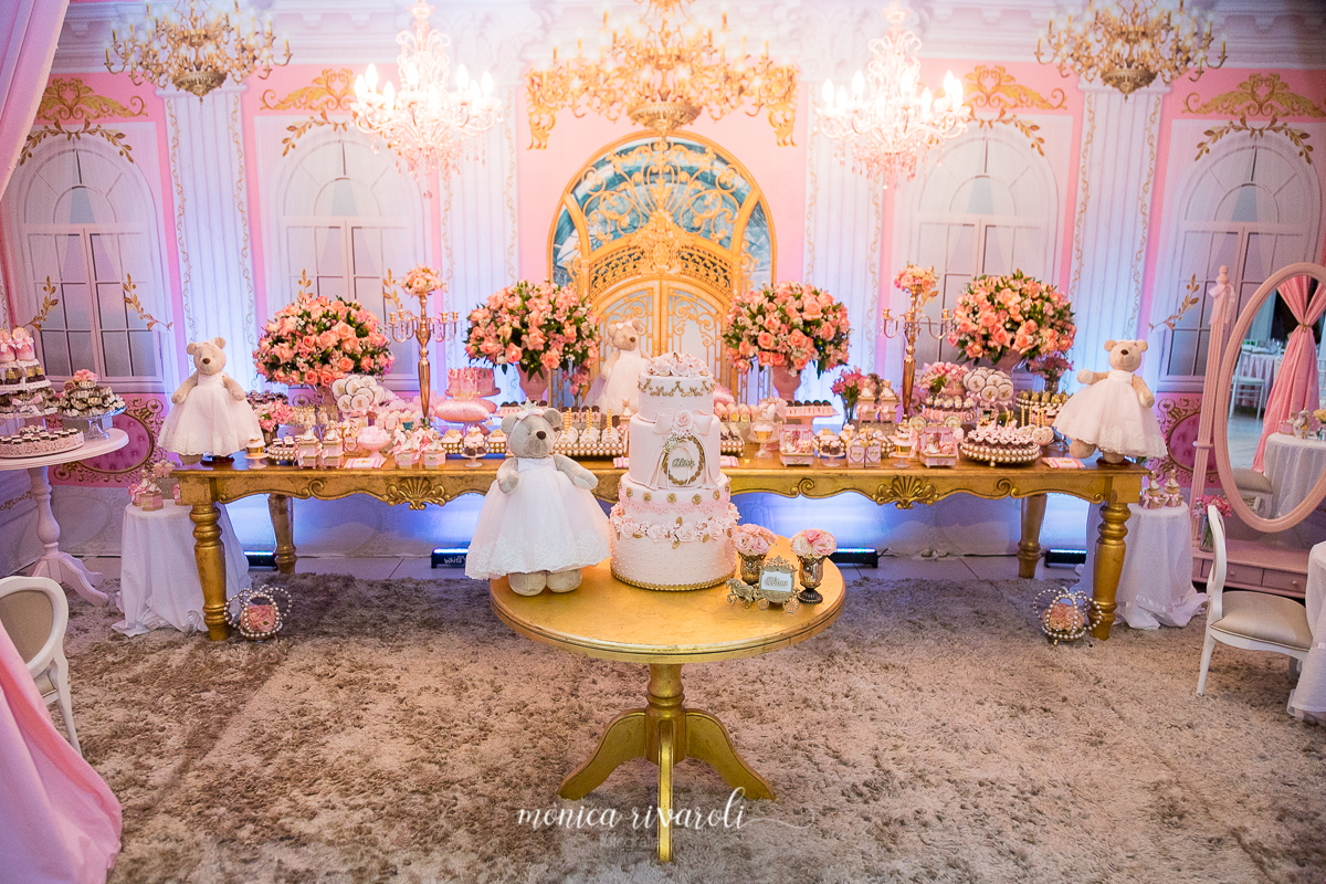 A mesa principal com o bolo tem um ursa vestida de princesa e com coroa em tons de bege, com a roupa rosa. Toda decoração tem detalhes em dourado e rosa.