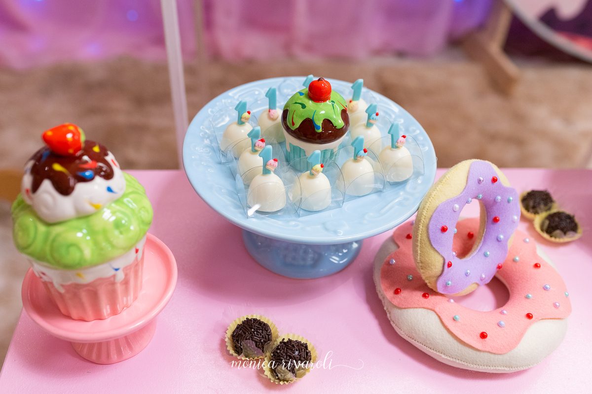 Detalhes da mesa de doces. Nesta foto, há um aparador com bombons em número 1 em cima, dois donnuts de feltro e um aparador com um cupcake de porcelana