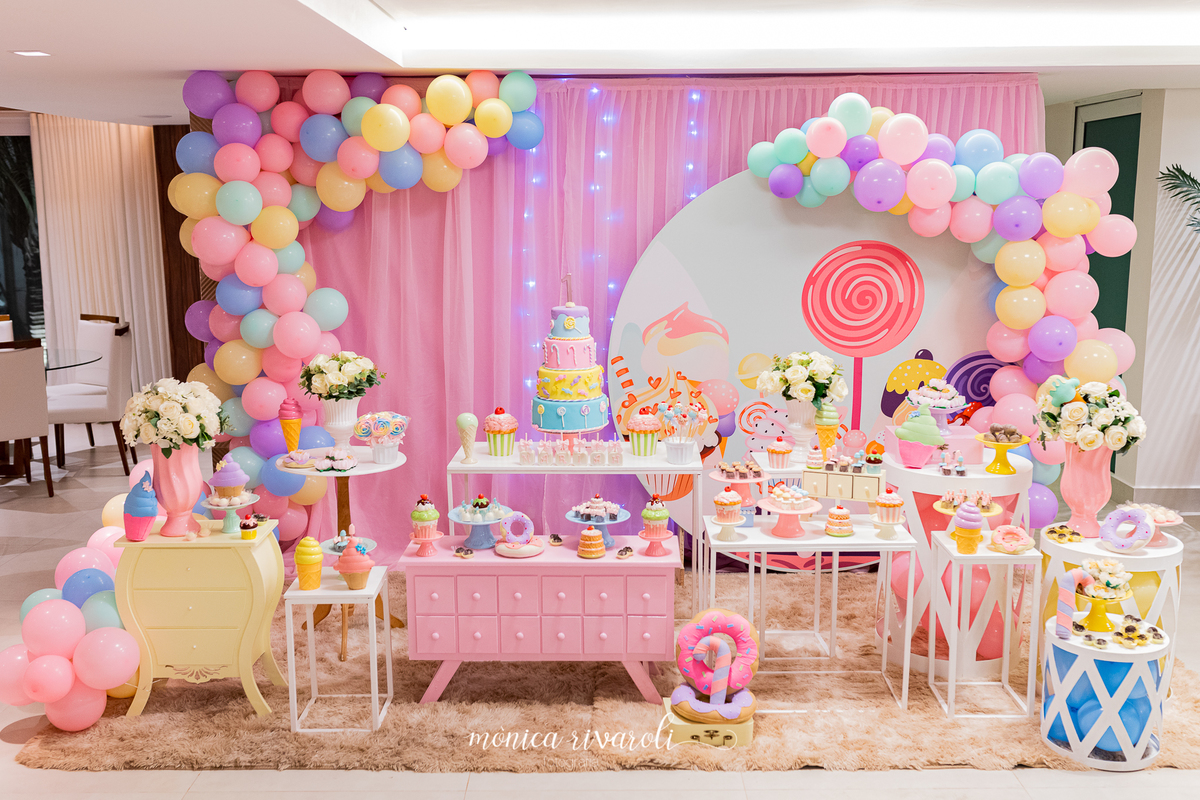 Fotografia da decoração da festa. O tema é doce. Nas mesas, tem vários aparadores com docinhos. Há também um arco de bal~oes