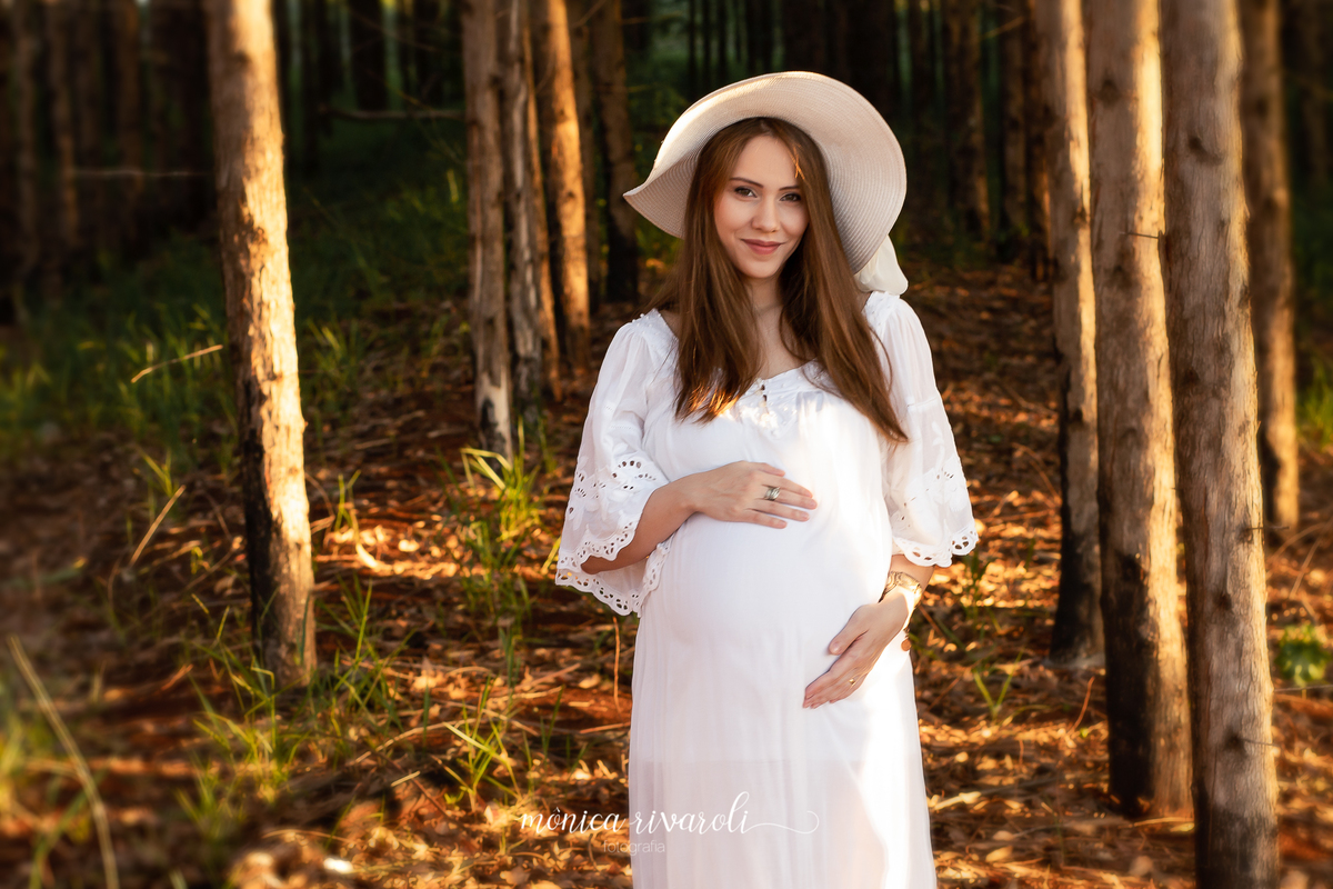 Em uma plantação de eucalipto, a gestante está com um vestido branco e um chapéu da mesma cor. Ela está com as mãos na barriga e sorrindo.
Fotografado por Mônicas Fotografia em Maringá-PR