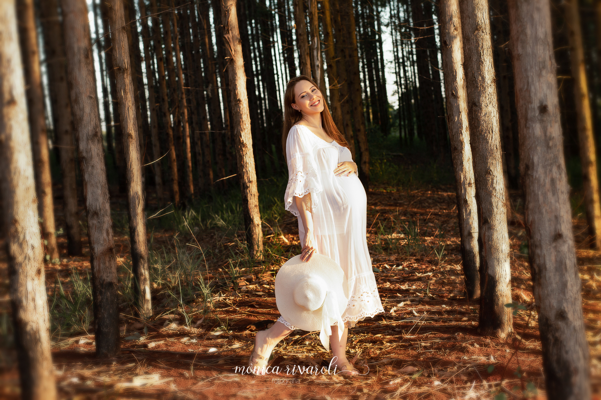 Em uma plantação de eucaliptos, a gestante está com uma mão na barriga e a outra mão está segurando um chapéu. Um dos pés está para trás e ela está sorrindo.
Fotografado por Mônicas Fotografia em Maringá-PR