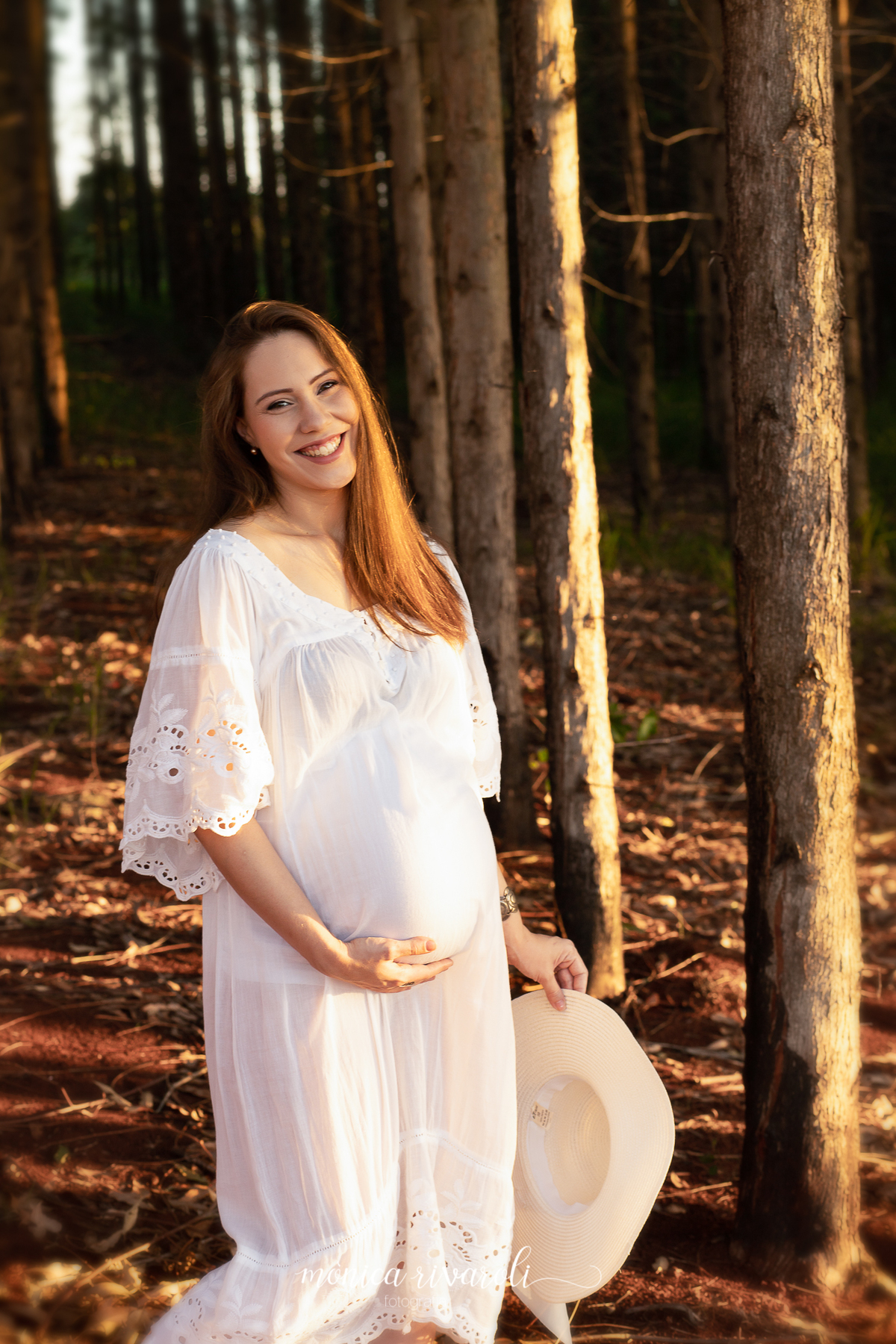 Ainda no mesmo cenário, a mãe grávida está olhando para a foto sorrindo. Uma mão está na barriga e a outra está segurando um chapéu branco.
Fotografado por Mônicas Fotografia em Maringá-PR