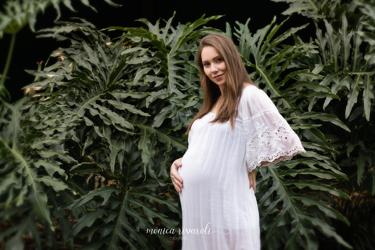 Em um fundo com plantas, a mãe grávida está com uma mão na barriga e a outra nas costas. A cabeça está levemente inclinada para o lado.
Fotografado por Mônicas Fotografia em Maringá-PR
