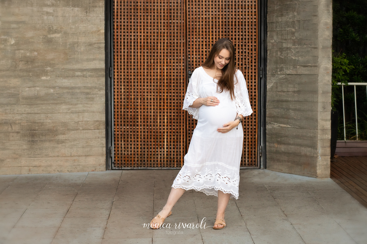 Em frente a uma parede com uma porta marrom, a gestante está com as duas mãos na barriga e com os pés mais afastados. Ela está olhando para a barriga e sorrindo.
Fotografado por Mônicas Fotografia em Maringá-PR