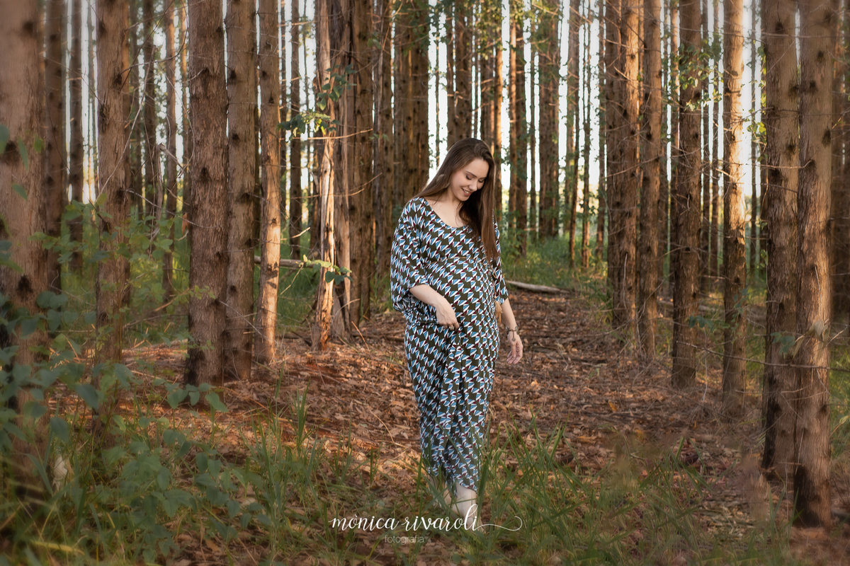 A mãe grávida está andando no meio das árvores, segurando o vestido e olhando para baixo com um sorriso
Fotografado por Mônicas Fotografia em Maringá-PR