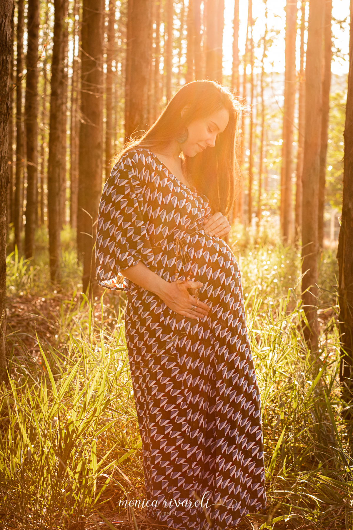 Em um lugar cheio de árvores, a mamãe está olhando para a Luisa com as mãos na barriga. A foto está bem iluminada pelo sol.
Fotografado por Mônicas Fotografia em Maringá-PR