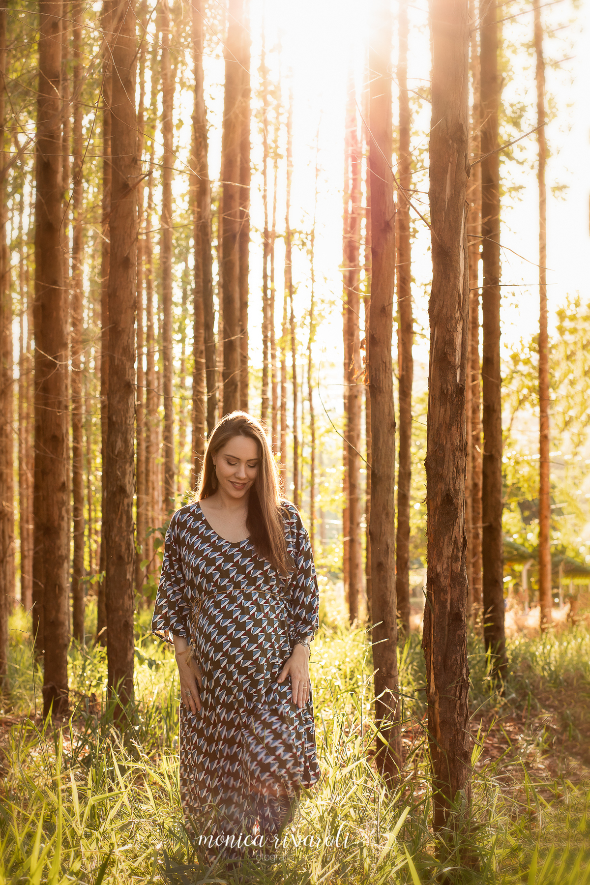 A gestante está caminhando no meio das árvores com o sol iluminando a foto. Ela está sorrindo e olhando para baixo.
Fotografado por Mônicas Fotografia em Maringá-PR