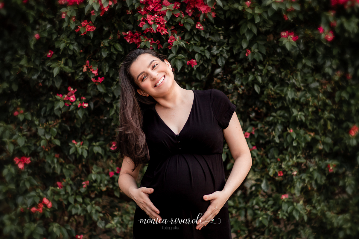 Em frente a um paredão de folhas e flores magenta, a Kelly está ao meio, com as duas mãos segurando a barriga, enquanto faz o movimento de jogar com o cabelo, ela sorri para a câmera. 
