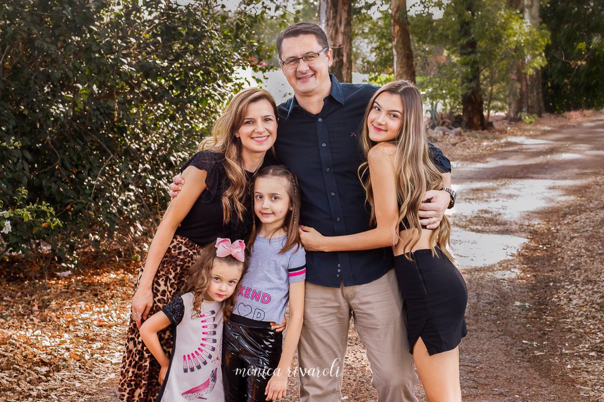 Nessa fotografia a família, o pai, a mãe e as três filhas, se abraçam em uma estrada de chão rodeada por árvores. A fotografia tem tons terrosos, deixando as árvores com tons de verdes mais opacos. 