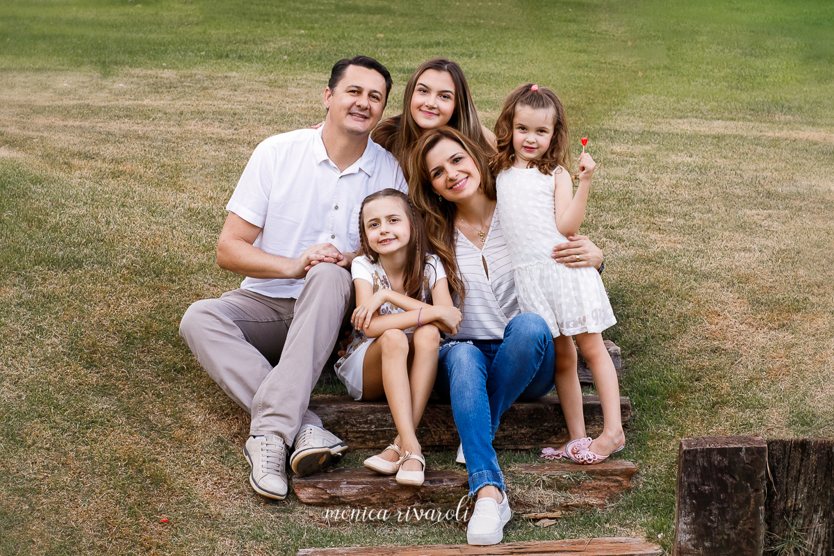 A família toda está sentada em uma escada de madeira embutida na grama. O pai na esquerda, no degrau debaixo a filha do meio, ao lado da mãe que está com o braço envolta da caçula, que segura um pirulito na mão. E atrás, no ultimo degrau, a mais velha. 