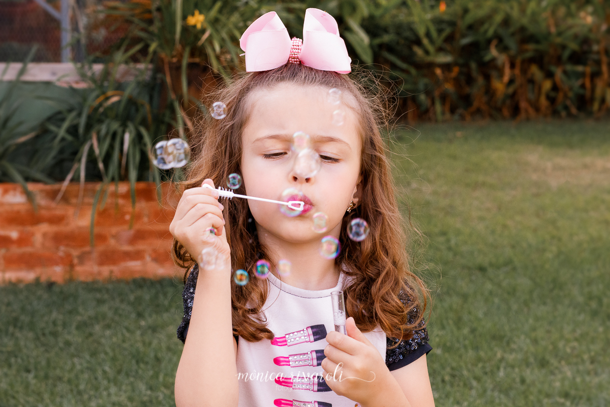 Nesta fotografia, a esquerda temos um murro de tijolos com flores em cima e na direita grama. Ao meio da foto a irmã caçula assopra e faz bolhas, ela usa um laço rosa sobre a cabeça prendendo só a franja do cabelo enrolado. 