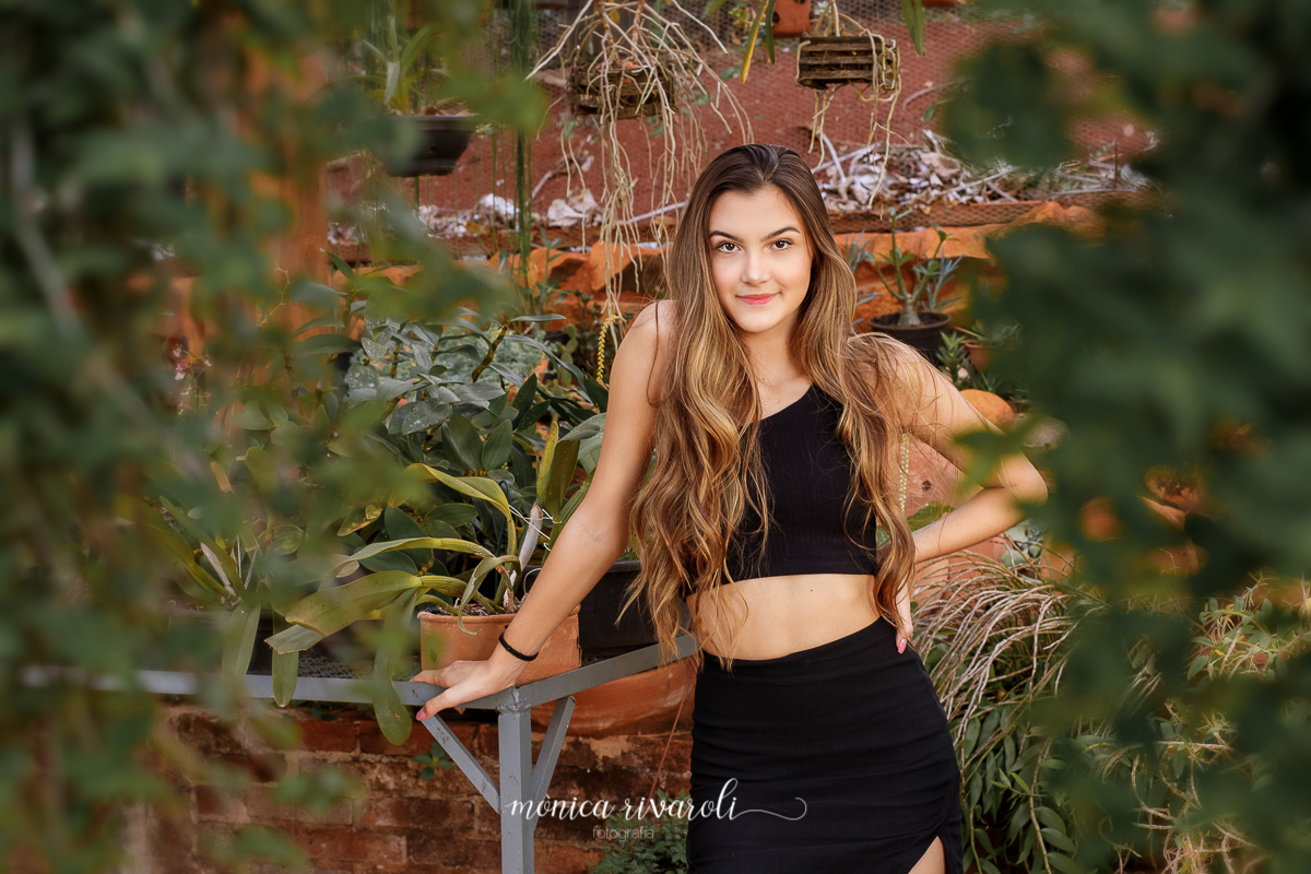 Nesta fotografia, a irmã mais velha está dentro da estufa, nos cantos inferiores da fotografia tem flores em tons de verde escuro, ao centro a menina apoia uma mão na mesa de metal, com vasos de flores, e a outra na cintura. O cabelo ondulado está de lado