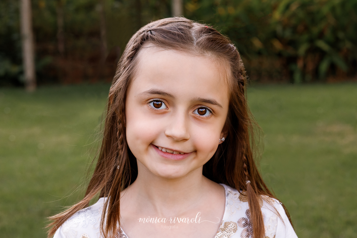O plano de fundo dessa fotografia e uma grama em tons terrosos e uma parede repleta de plantas. Ao meio a irmã caçula sorri para foto. Ela usa um vestido branco e o cabelo liso esta repartido ao meio. A franja esta presa em duas tranças, uma de cada lado.