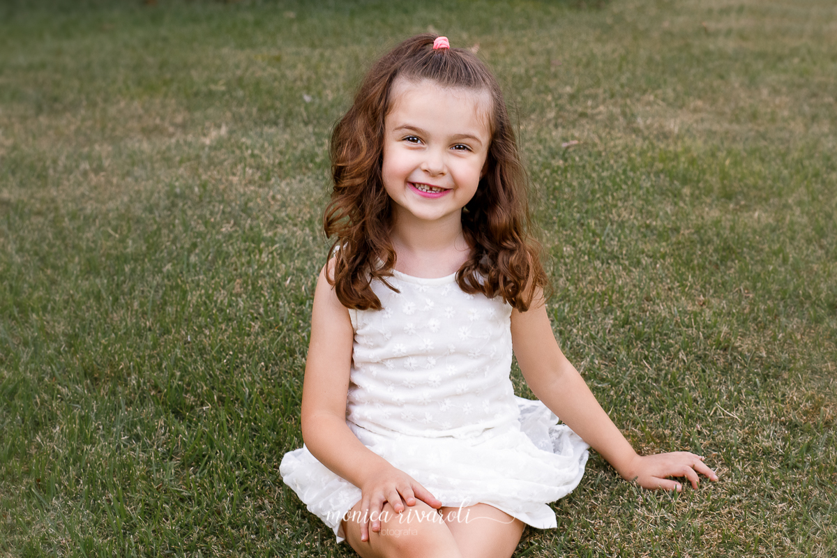 Nesta fotografia a irmã caçula esta sentada na grama, ela sorri para foto usando um vestido branco, suas pernas estão cruzadas na altura do joelho. Sua está presa em 