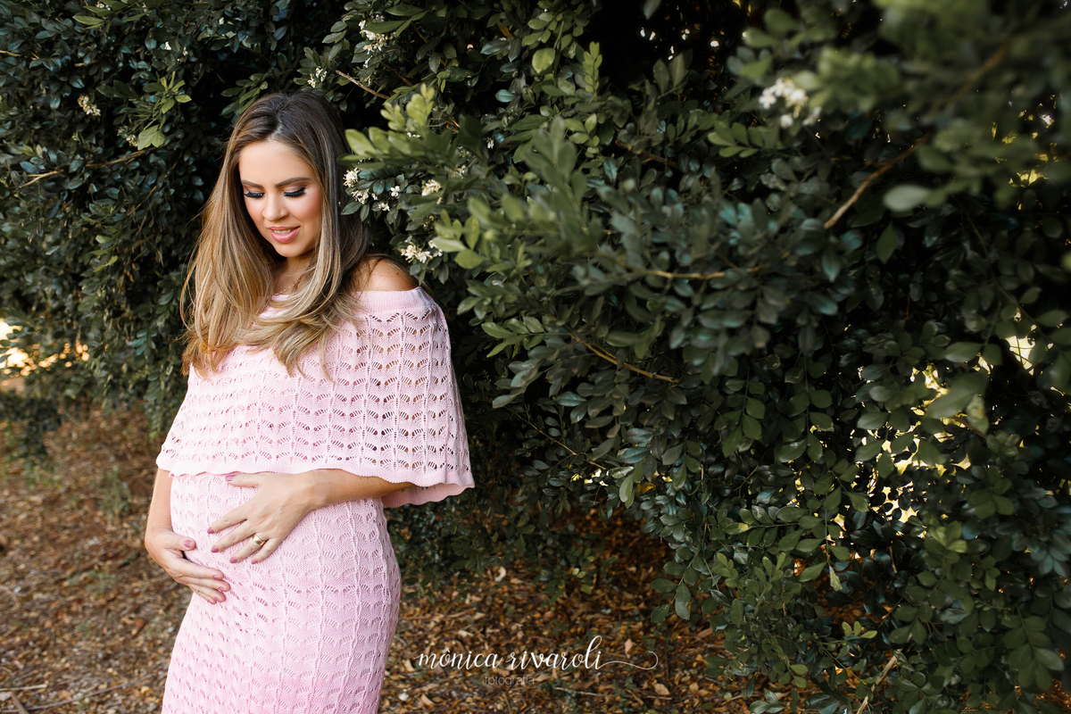 A mãe grávida está ao lado de uma parede de arbustos, ela está com um vestido rosa e as mãos na barriga. A mamãe está olhando para o bebê na barriga com um sorriso.
Ensaio Gestante
Fotografado por Mônicas Fotografia em Maringá-PR