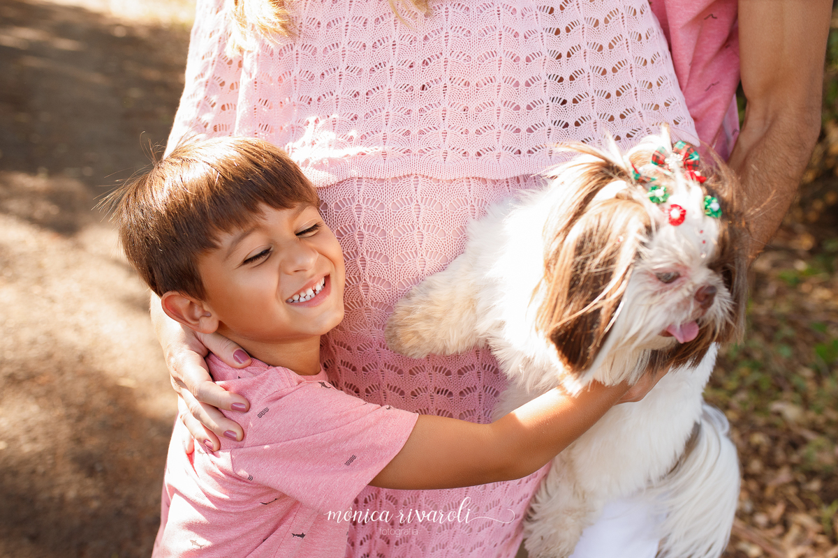 O irmão mais velho está abraçando a mãe e sorrindo com os olhos fechados. A gestante está com uma mão no filho e com a outra está segurando a cachorrinha da família. Na foto aparece apenas a barriga da mãe.
Fotografado por Mônicas Fotografia em Maringá-PR