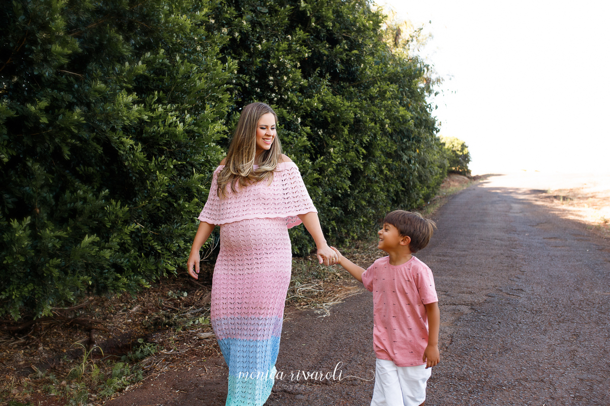 Em uma rua com um parede de arbustos no canto, a mãe grávida está caminhando com o filho mais velho. Eles estão de mãos dadas e sorrindo um para o outro.
Ensaio Gestante
Fotografado por Mônicas Fotografia em Maringá-PR