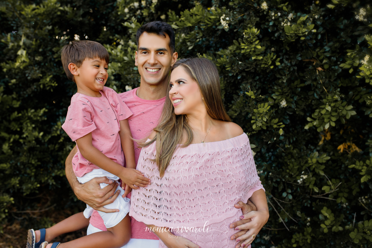 Em uma parede de arbustos, o pai está com o filho mais velho no colo e com o outro braço abraça 
 a gestante. A mãe está com uma mão na barriga e olhando para o filho. O irmão mais velho está rindo.
Fotografado por Mônicas Fotografia em Maringá-PR
