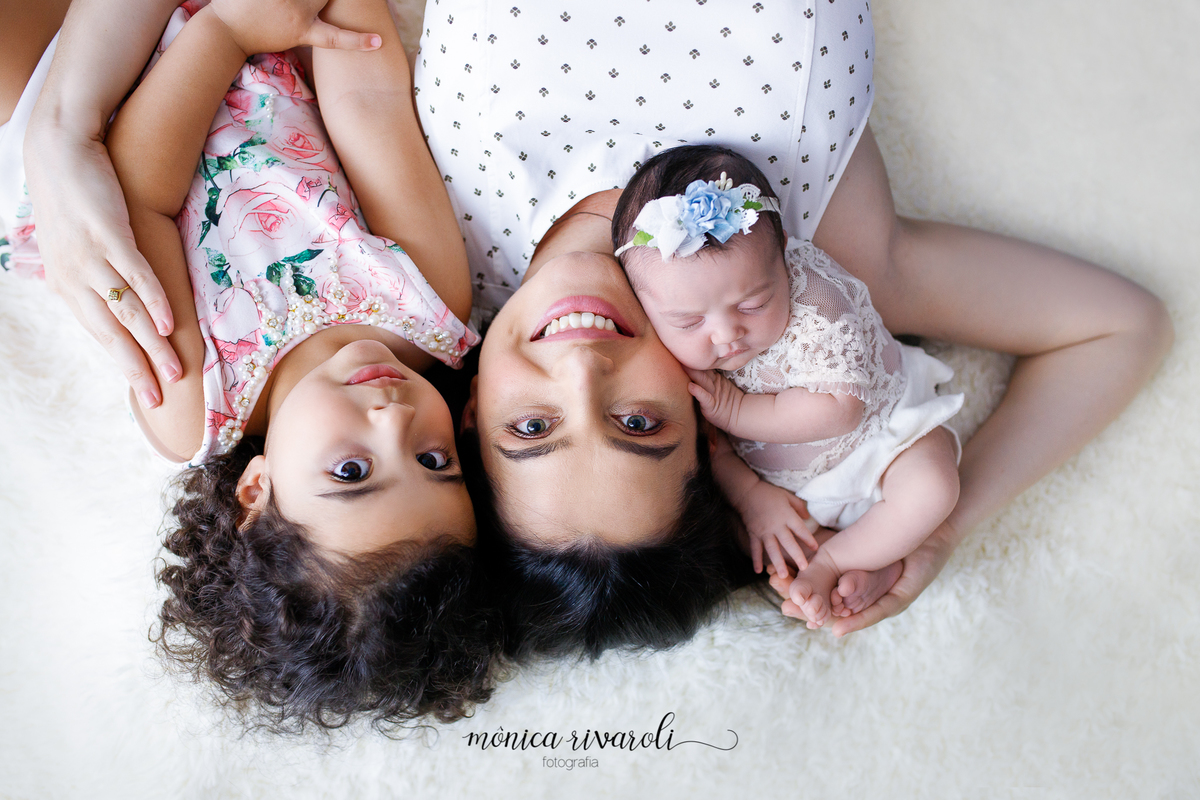 A mãe está deitada com as duas filhas em um fundo branco, ela está no meio. A mãe, a irmã mais velha e a bebê estão com os rostos próximos.
Ensaio newborn
Fotografado por Mônicas Fotografia em Maringá-PR