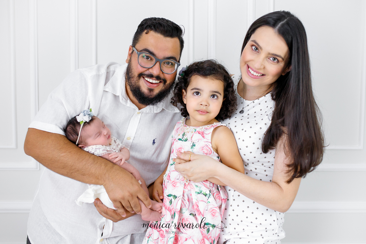 Em um cenário branco, a filha mais velha está entre a mãe e o pai que está segurando a bebê no colo. A mãe está segurando a mão da filha mais velha.
Ensaio Newborn
Fotografado por Mônicas Fotografia em Maringá-PR