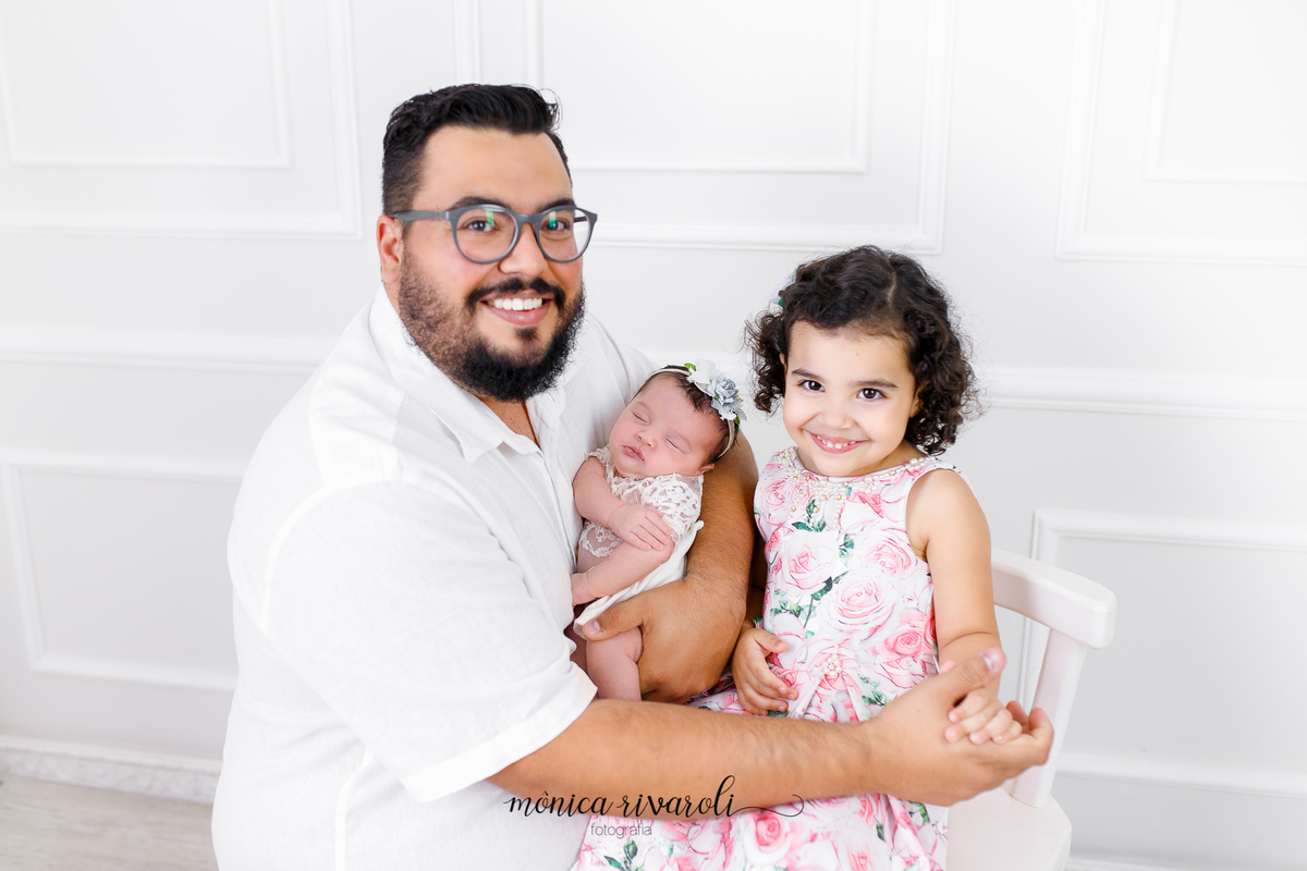 Em um cenário branco, a filha mais velha está sentada em uma cadeira e o pai está com a bebê no colo agachado ao lado. O pai está segurando a mão da filha mais velha, os dois estão sorrindo
Ensaio Newborn
Fotografado por Mônicas Fotografia em Maringá-PR
