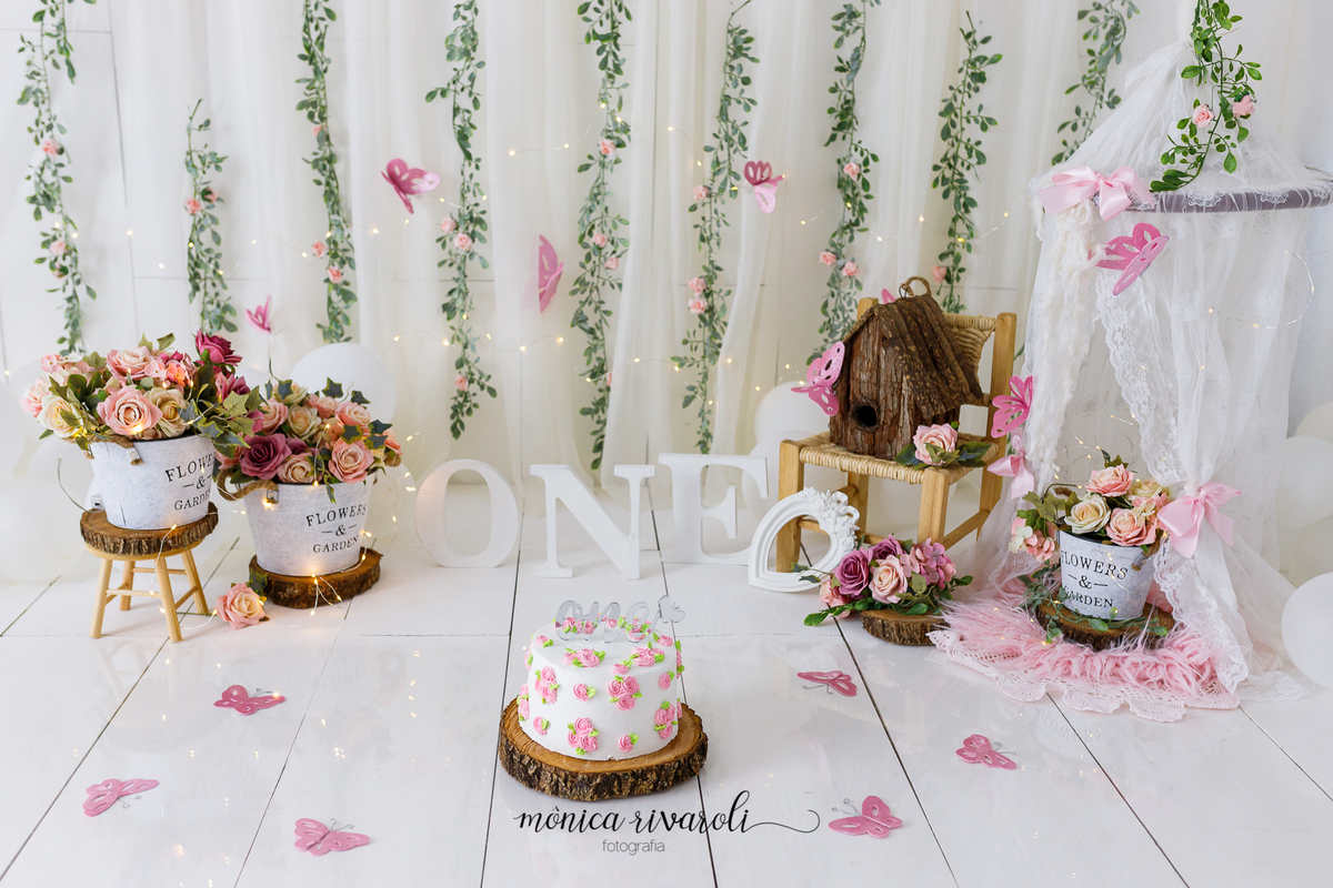 A foto mostra um cenário de smash the Cake, um bolo branco com flores rosa está no centro. No fundo há flores em vasos, plantas e borboletas de papel rosa. Também há letras formando a palavra