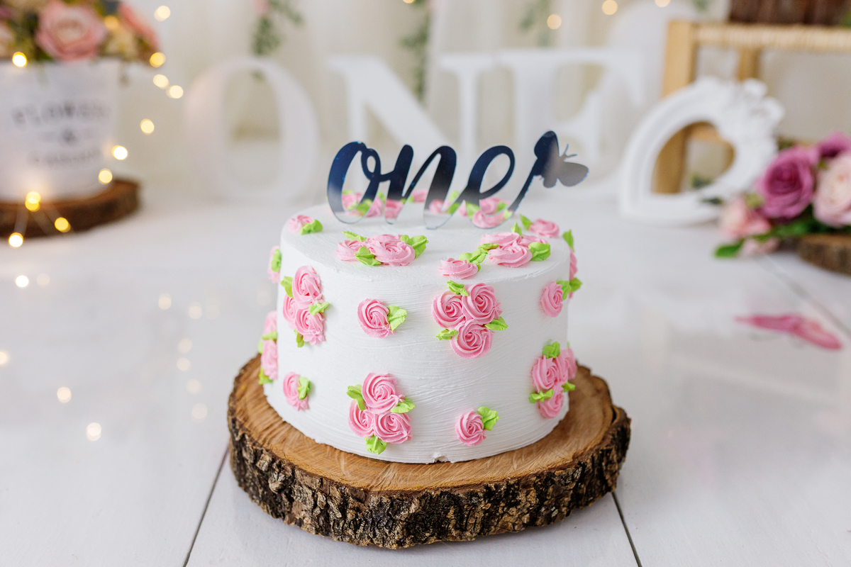 A foto mostra os detalhes do bolo do smash the cake. Um bolo branco com flores rosa espalhadas, no topo há a palavra