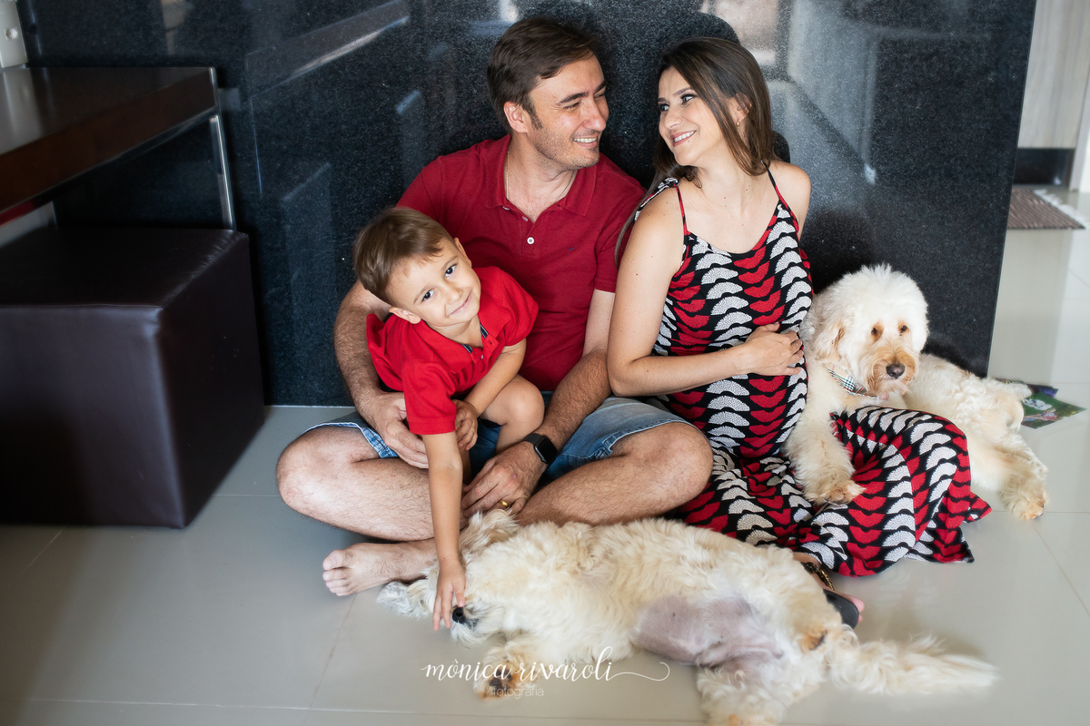 A mamãe e o pai estão sentados no chão da sala com o filho mais velho no colo do pai e os cachorros da família estão com eles. Os pais estão se olhando e sorrindo.
Fotografado por Mônicas Fotografia em Maringá-PR