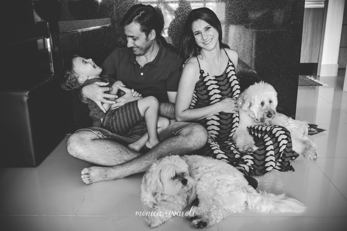 O filho mais velho está no colo do pai rindo e o pai está olhando para ele sorrindo. A mãe grávida está sorrindo e olhando para frente com a mão na barriga
A foto está em preto e branco
Fotografado por Mônicas Fotografia em Maringá-PR