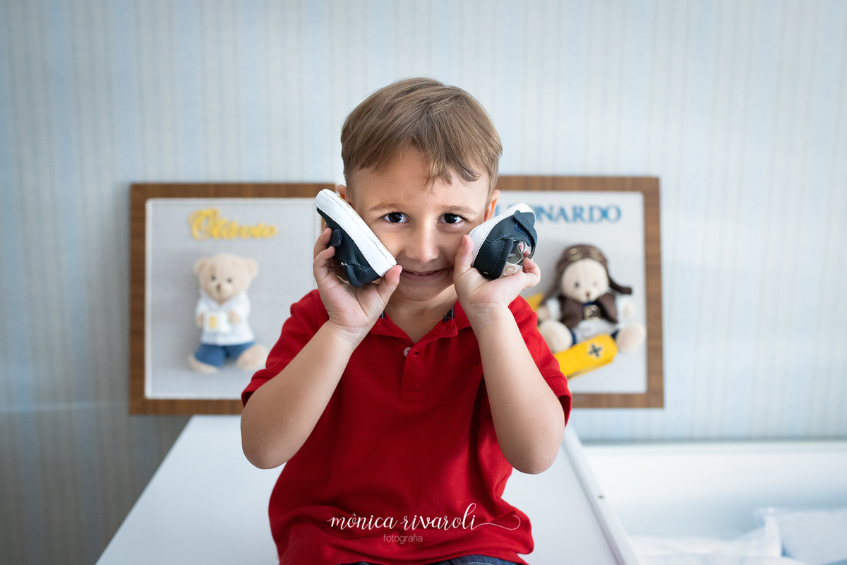 No fundo tem dois quadros, um com o nome Leonardo e o outro com o nome Otávio. O filho mais velho está sorrindo com os sapatinhos do irmão mais novo perto do rosto
Fotografado por Mônicas Fotografia em Maringá-PR