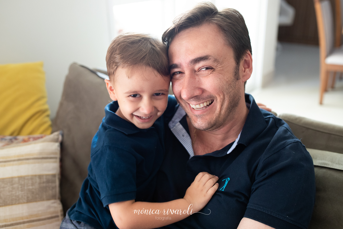 Ainda no sofá, o filho mais velho está sentado no colo do pai e os dois estão sorrindo
Fotografado por Mônicas Fotografia em Maringá-PR