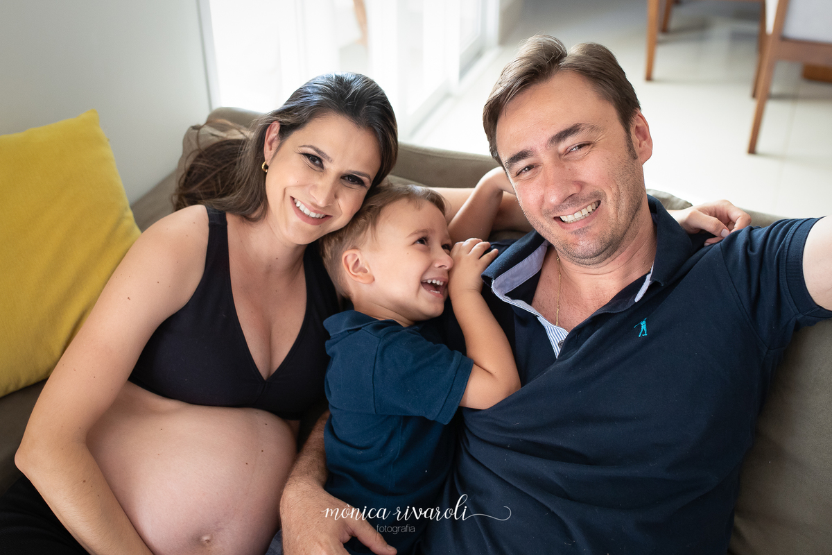 A mãe grávida, o pai e o filho mais velho estão sentados no sofá. O filho mais velho está no meio dos dois olhando pro pai e todos estão sorrindo
Fotografado por Mônicas Fotografia em Maringá-PR
