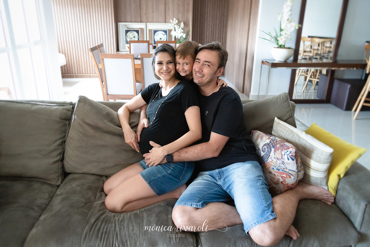 Mãe grávida, pai e irmão mais velho sentados no sofá de casa. O irmão está atrás dos pais com o rosto no meio deles. Estão todos abraçados.
Fotografado por Mônicas Fotografia em Maringá-PR