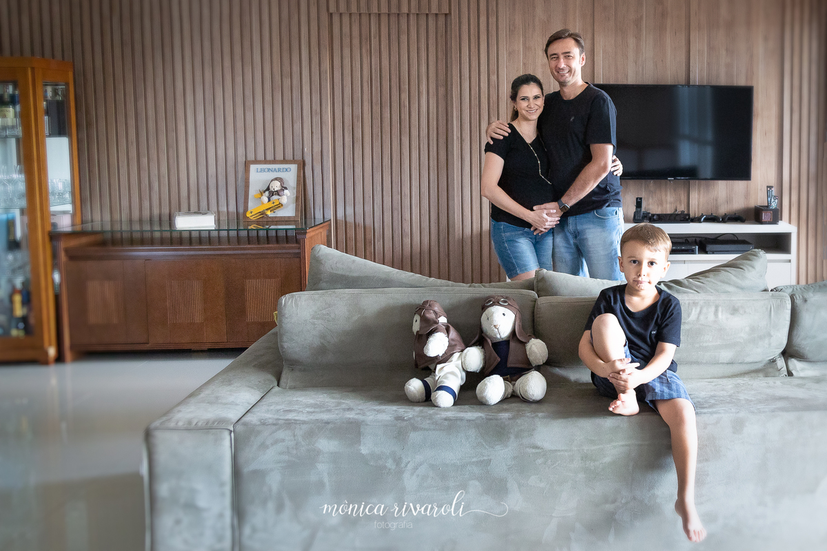 Filho mais velho, chamado Otávio, está sentado no sofá da sala do lado de dois brinquedos de pelúcia e atrás do sofá a mamãe e o papai estão sorrindo.
Fotografado por Mônicas Fotografia em Maringá-PR