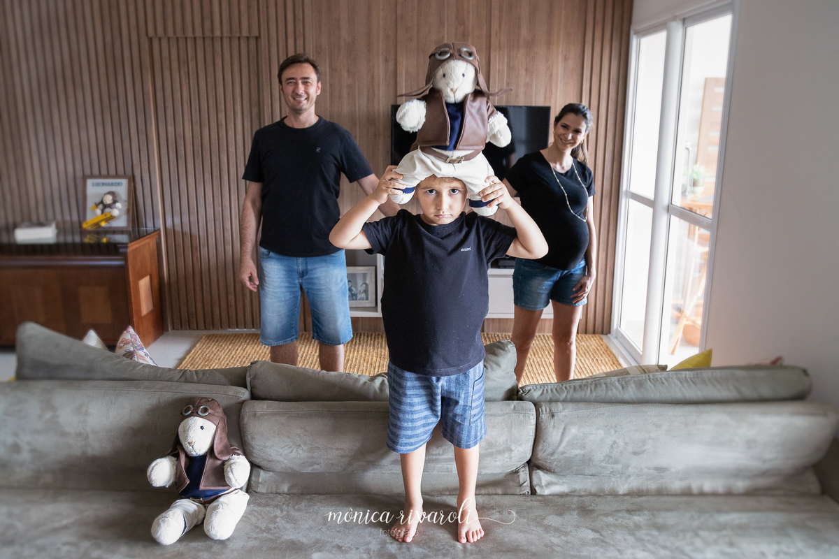 O filho mais velho está em pé no sofá com um brinquedo de pelúcia na cabeça. Atrás do sofá estão os pais sorrindo.
Fotografado por Mônicas Fotografia em Maringá-PR