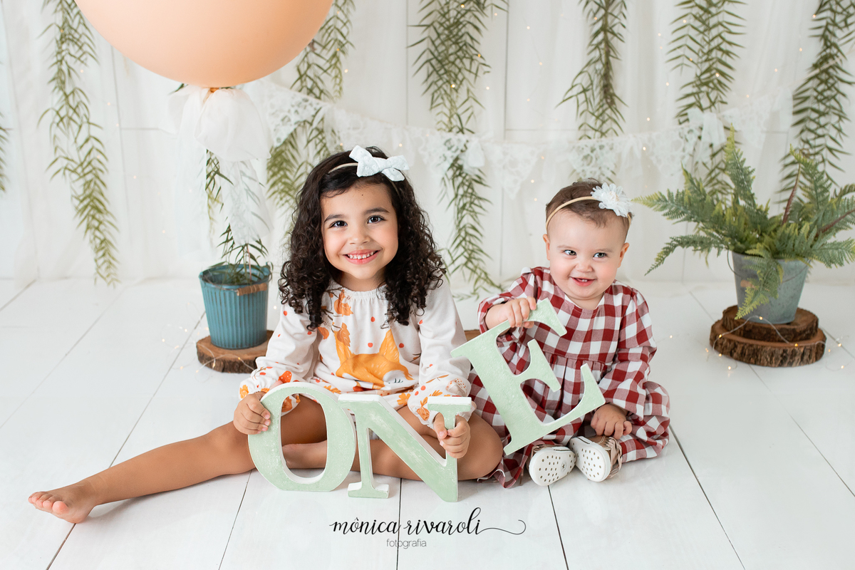 No cenário de Smash The Cake, a bebê está sentada ao lado da sua irmã mais velha. As duas estão sorrindo e segurando as letras da palavra 