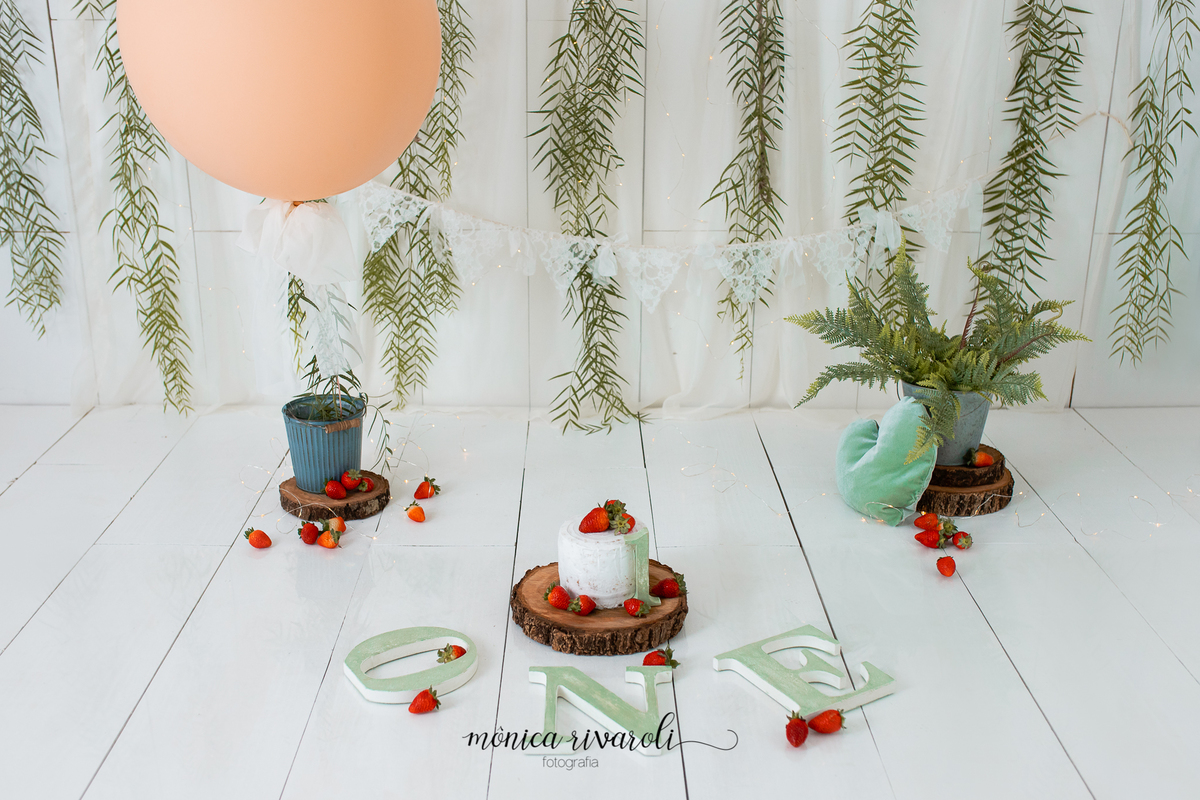 A foto mostra o cenário do Smash The Cake. No centro está em um bolo com morangos, na frente as letras formando a palavra 