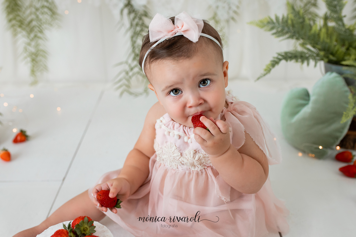 No cenário de Smash, a bebê com um vestido rosa está comendo um morango e na outra mão está segurando outro morango
Ensaio Smash The Cake
Fotografado por Mônicas Fotografia em Maringá-PR
