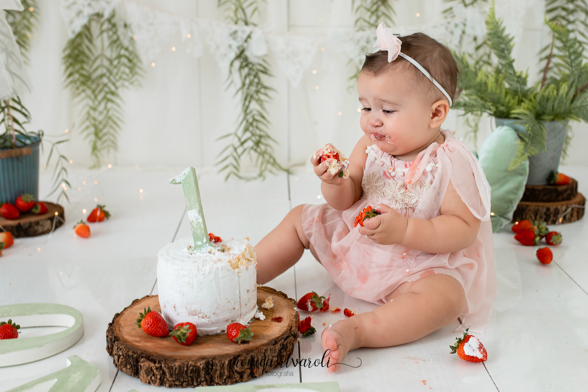 A bebê está olhando para o morango na sua mão e fazendo careta. No cenário tem um bolo, plantas e morangos espalhados.
Ensaio Smash The Cake
Fotografado por Mônicas Fotografia em Maringá-PR