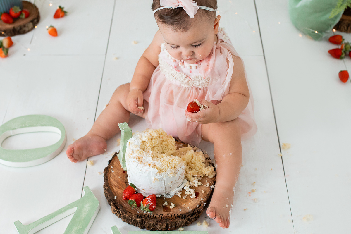 A foto foi tirada de cima para baixo, a bebê está olhando para o morango na sua mão. Na frente dela está o bolo que a menina brincou
Ensaio Smash The Cake
Fotografado por Mônicas Fotografia em Maringá-PR
