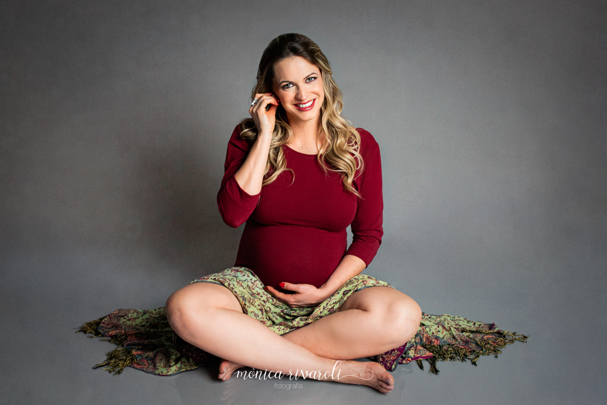 Num fundo cinza, a mamãe está sentada no chão, em posição de indiozinho, com um body de mangas longas vermelho e uma manta colorida sobre o colo. Ela sorri para a foto, com uma mão embaixo da barriga e a outra na orelha. 