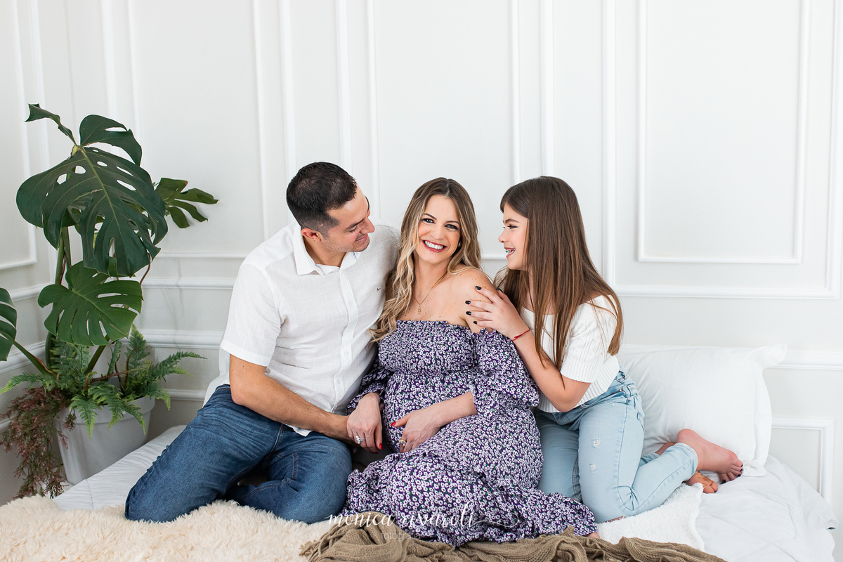 Ainda no mesmo cenário, a mamãe olha para a câmera enquanto o papai e a filha a olham. Eles estão sorrindo. 