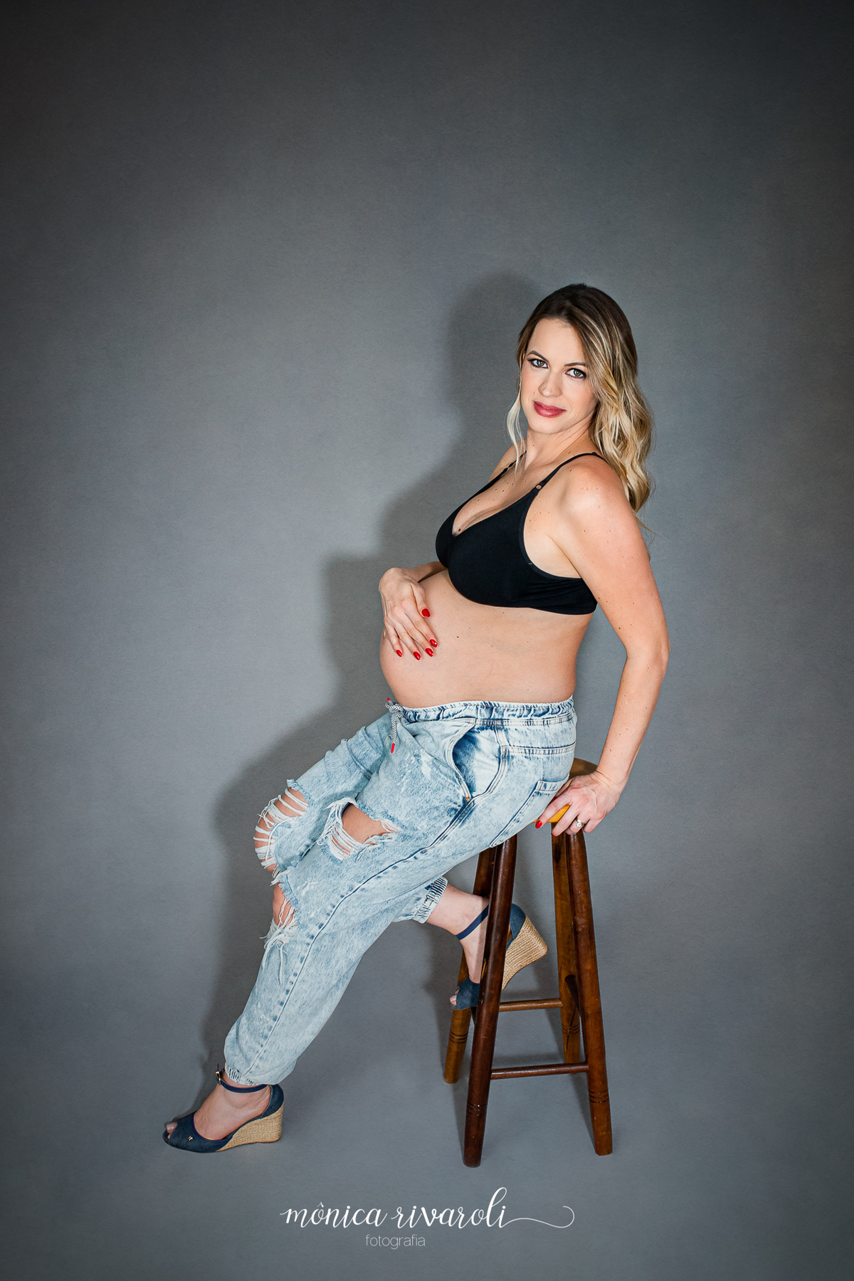 Agora a mamãe está sentada em um banco alto de madeira, ela usa uma calça jeans rasgada e um top preto. Uma das suas mãos está apoiada no banquinho e a outra na barriga. Ela olha para a câmera.